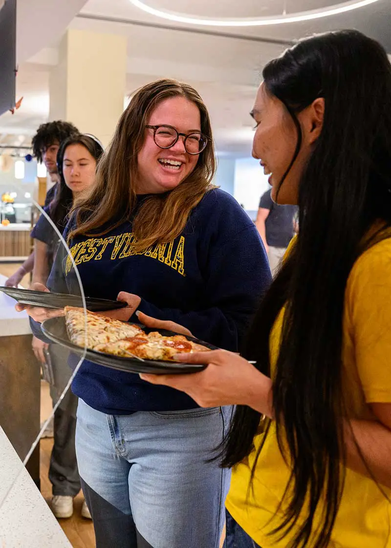 Two students holding dishes and smiling while standing in cafeteria
